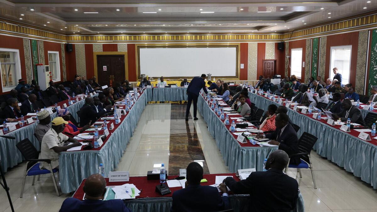 Large conference room with participants seated around a U-shaped arrangement of tables, facing a speaker at the center, with nameplates and documents on the tables.