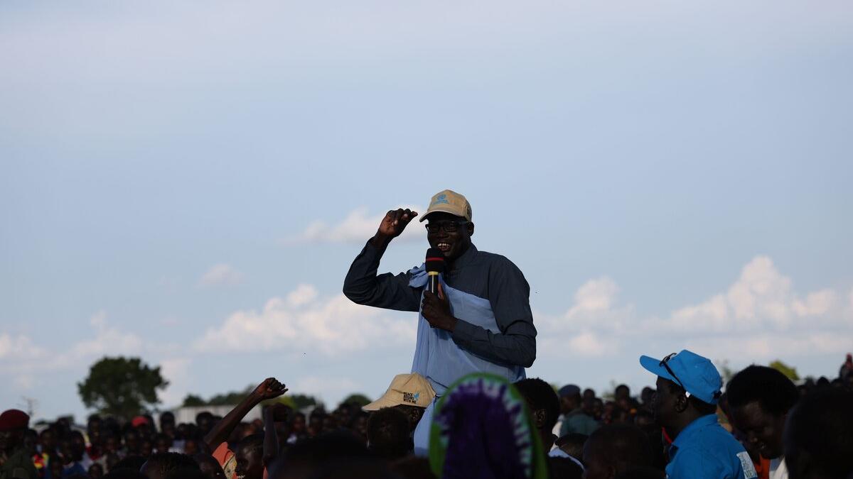 Person holding a microphone and papers while addressing a large outdoor crowd under a clear sky.
