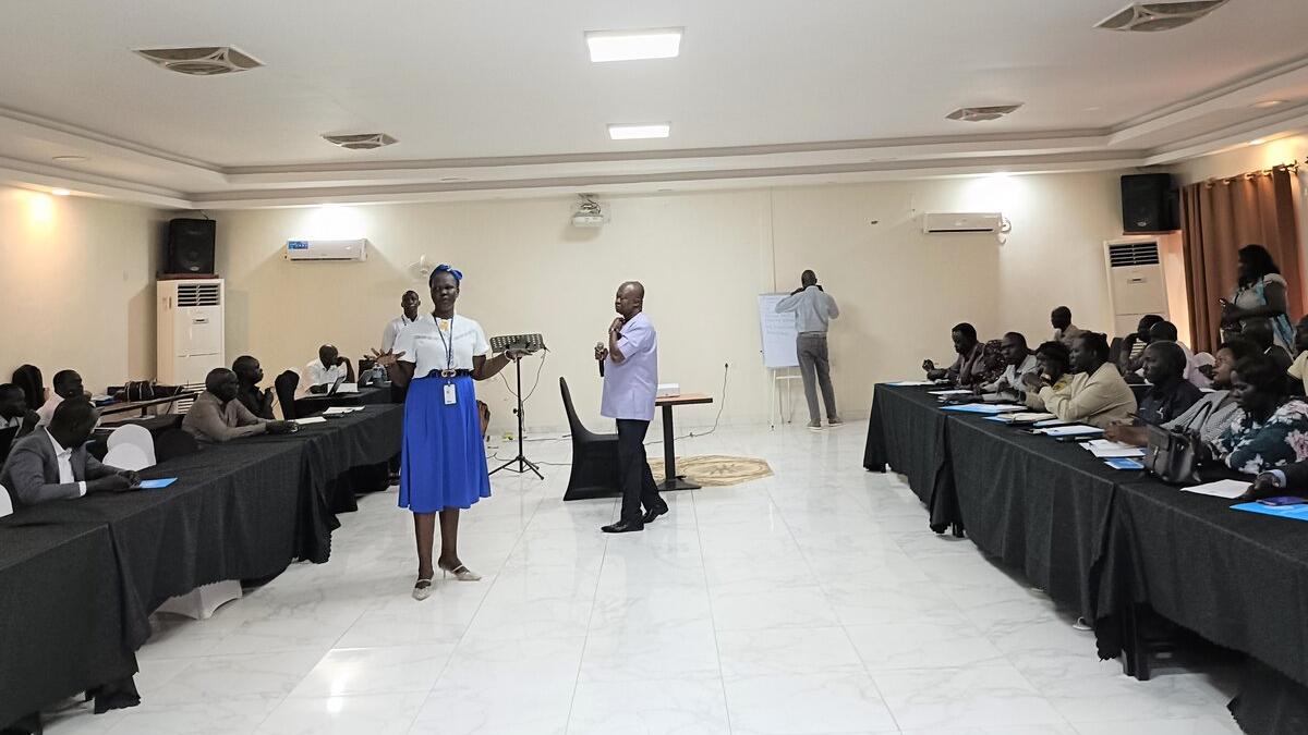 A training session in a conference room with two facilitators standing at the front, participants seated at long tables, and a person writing on a flip chart in the background