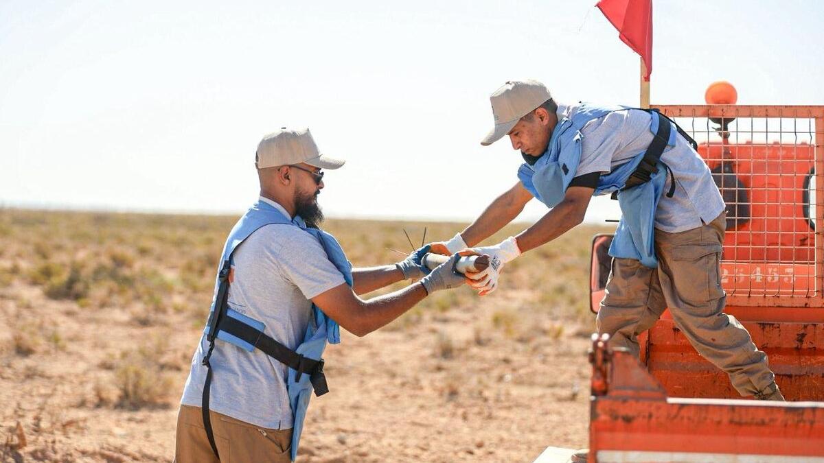 A high-angle shot looking down into a shallow, dug-out pit in dry, rocky desert soil. Inside the pit, approximately five to six large, rusted cylindrical munitions (likely large-caliber artillery shells or rockets) are carefully laid out in a row. They are resting on top of white and yellow sandbags, likely placed there to stabilize the ordnance or prepare it for controlled demolition or safe transport. The surrounding area is barren desert scrubland.