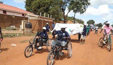People riding hand-powered tricycles lead a street parade, followed by others carrying a large banner, on a dirt road with buildings and trees in the background.
