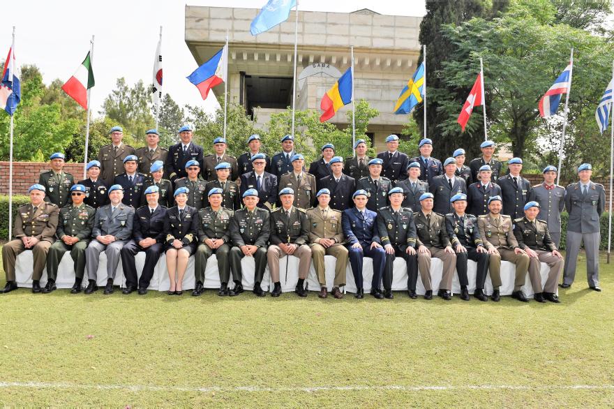 Group photo of UN military personnel in uniform and blue berets seated and standing in rows outdoors with national flags behind them.